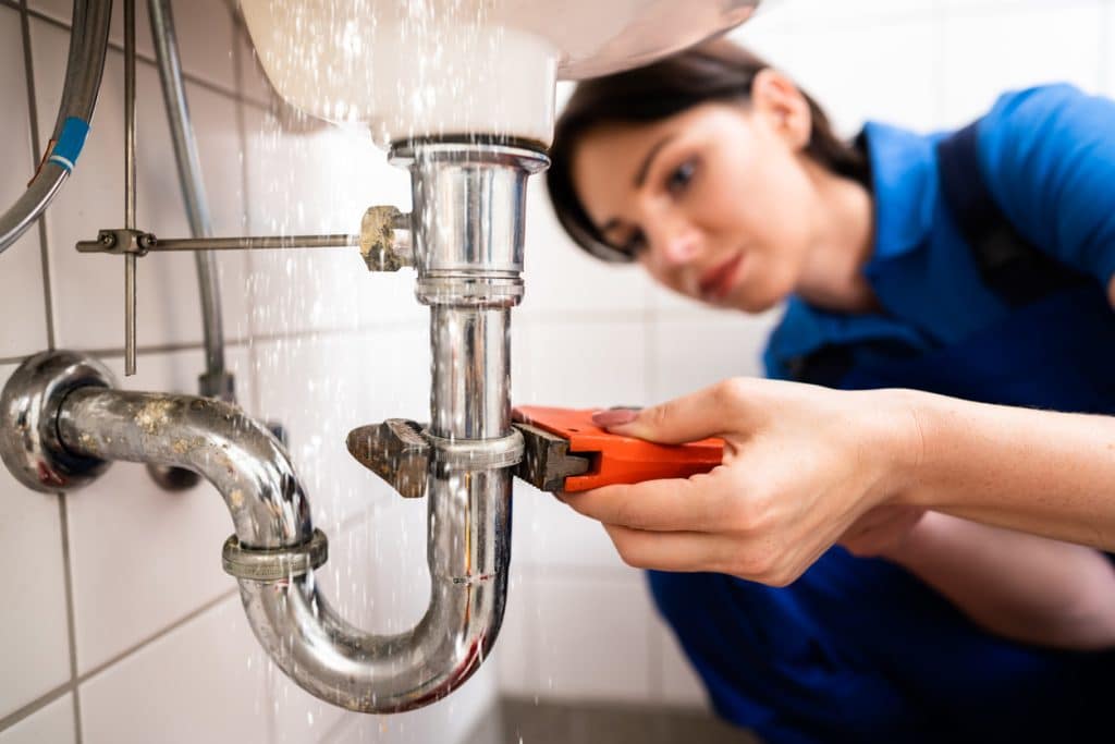 Plumber using a pipe wrench to repair a leaking sink drain pipe beneath a faucet.