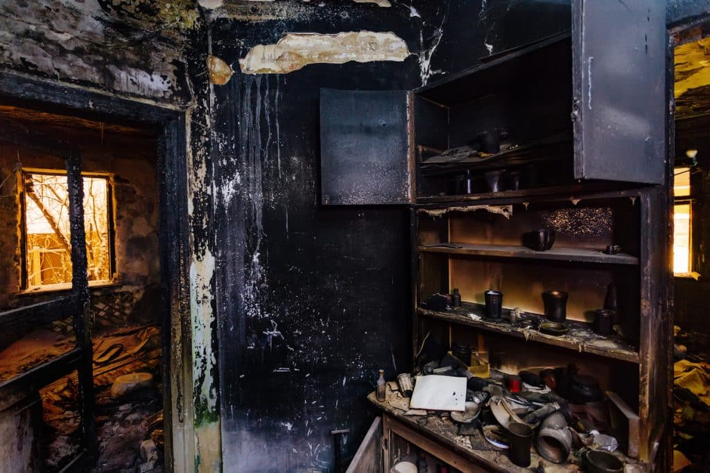 A severely fire-damaged house interior with blackened walls, charred shelving, damaged furniture, and debris scattered throughout the rooms.