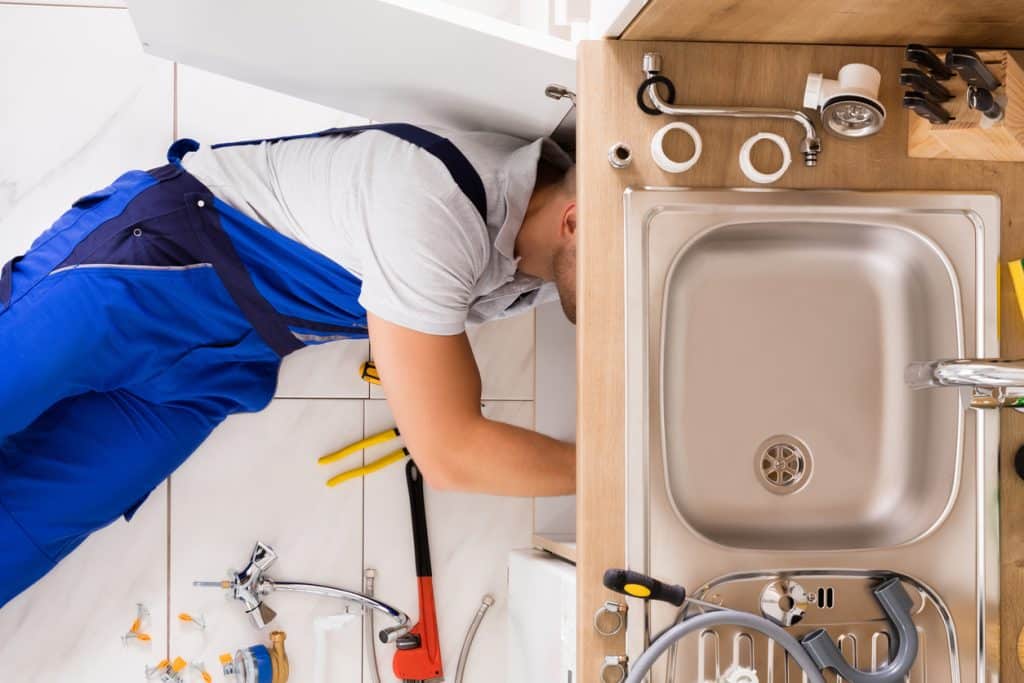 Plumber inspecting and repairing the plumbing beneath a stainless steel kitchen sink, with tools and replacement fittings nearby.